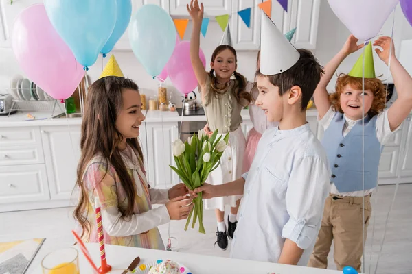 Menino feliz dando tulipas para menina de aniversário alegre perto de amigos no fundo borrado — Fotografia de Stock