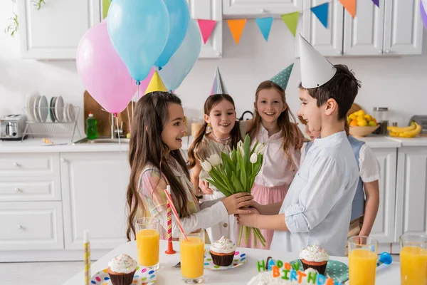 Happy preteen boy giving flowers to cheerful birthday girl near friends during party — Stock Photo