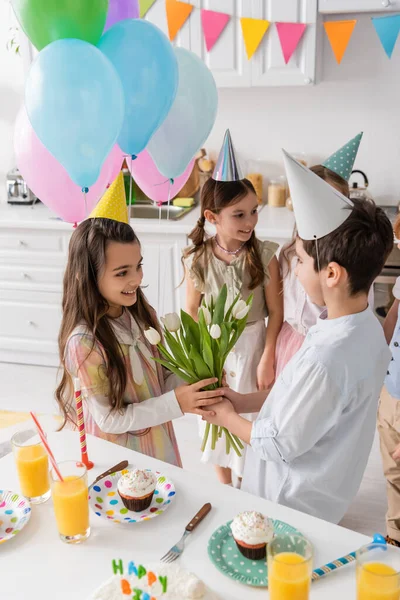Preadolescente chico dando tulipanes a alegre cumpleaños chica cerca de niños en fondo borroso - foto de stock