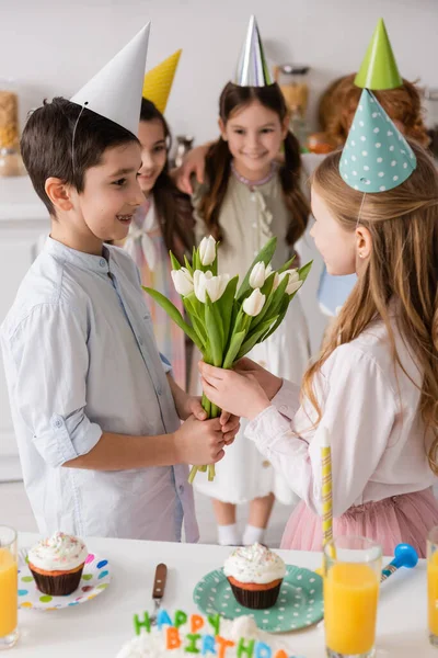 Menino pré-adolescente com aparelho dando tulipas para alegre aniversariante perto de amigos no fundo borrado — Fotografia de Stock