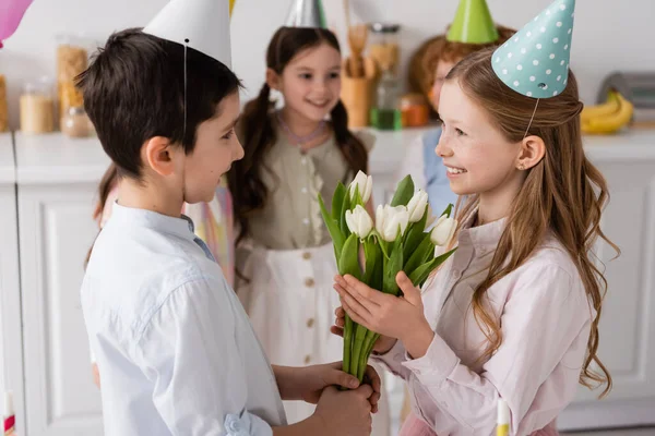 Позитивний хлопчик, що дарує тюльпани задоволеній дівчині на день народження біля друзів на розмитому фоні — стокове фото