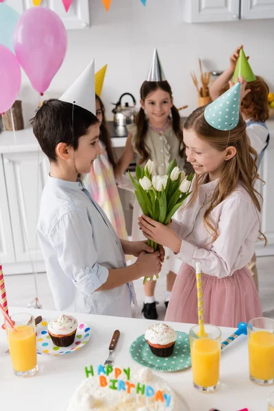 Preadolescente chico dando tulipanes a alegre cumpleaños chica cerca de cupcakes y amigos en fondo borroso - foto de stock