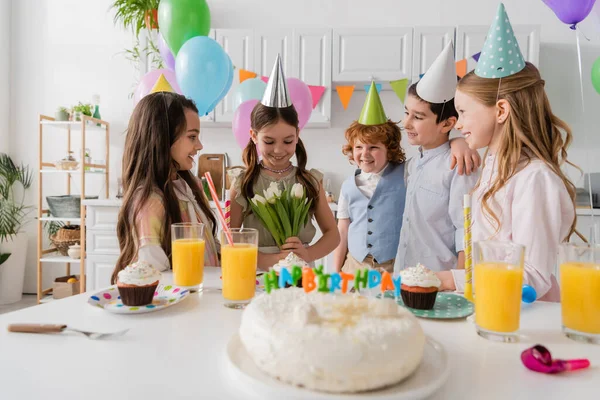 Cheerful birthday girl holding tulips near happy friends during celebration at home — Stock Photo