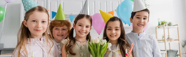 Alegre chica sosteniendo tulipanes cerca de amigos durante la fiesta de cumpleaños en casa, pancarta - foto de stock