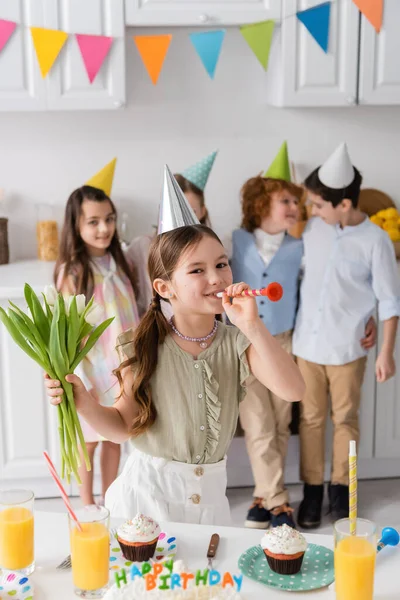 Alegre chica sosteniendo tulipanes y soplando cuerno de fiesta cerca de amigos durante la fiesta de cumpleaños en casa - foto de stock