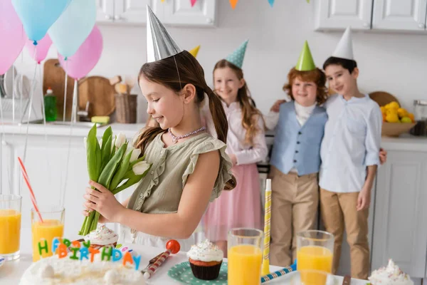Cheerful girl in party cap holding tulips next to friends during birthday party at home — Stock Photo