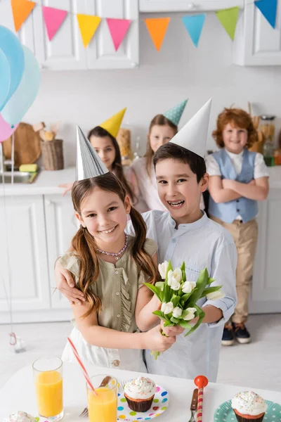 Menina positiva no boné do partido segurando tulipas perto do menino e ao lado de amigos durante a festa de aniversário em casa — Fotografia de Stock