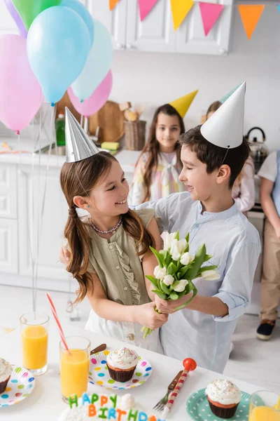 Happy girl in party cap holding tulips near boy and next to friends during birthday party at home — Stock Photo
