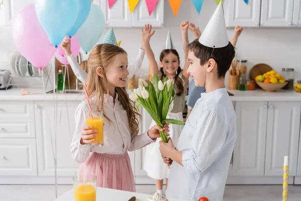 Glücklich Mädchen in Partymütze nimmt Tulpen von Jungen in der Nähe von Freunden auf verschwommenem Hintergrund — Stockfoto