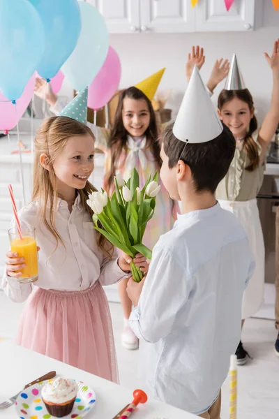Happy girl with glass of juice taking tulips from boy near friends on blurred background — Stock Photo