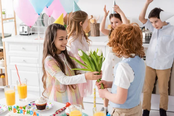 Happy girl in party cap taking tulips from redhead boy near friends on blurred background — Stock Photo