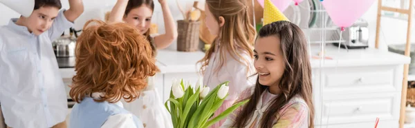 Glückliches Mädchen in Partymütze, das Tulpen von rothaarigen Jungen in der Nähe von Freunden auf verschwommenem Hintergrund nimmt, Banner — Stockfoto