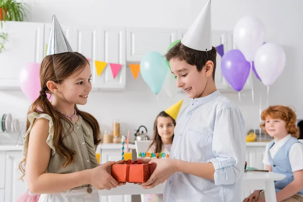 Щасливий хлопчик у дужках дарує подарунок веселій дівчині на день народження біля друзів на розмитому фоні — стокове фото
