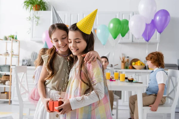 Cheerful birthday girl holding present and hugging friend during birthday party — Stock Photo