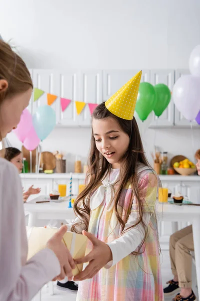 Amazed girl in party cap receiving birthday present from friend on blurred foreground — Stock Photo