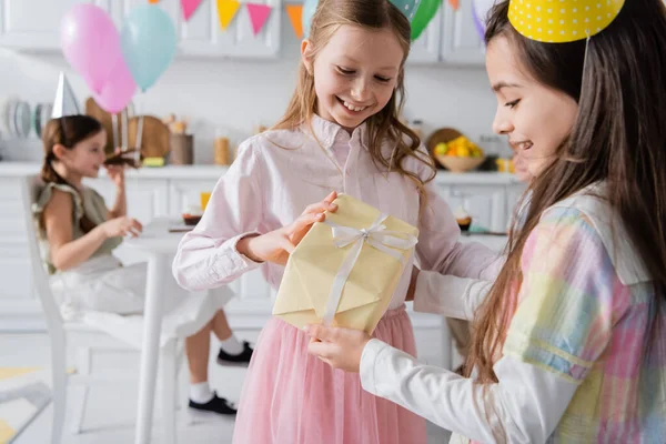 Cheerful birthday girl in party cap receiving present from happy friend — Stock Photo