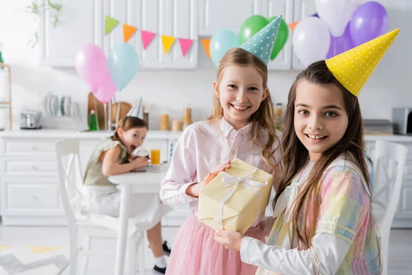 Menina de aniversário alegre em boné de festa recebendo presente de amigo enquanto olham juntos na câmera — Fotografia de Stock