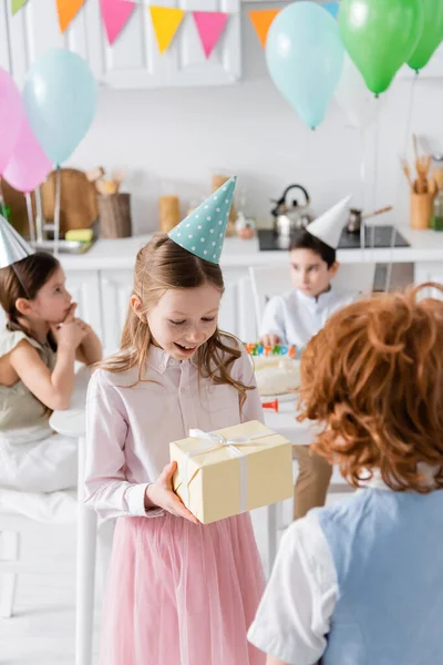 Amazed girl in party cap receiving birthday present from redhead boy — Stock Photo