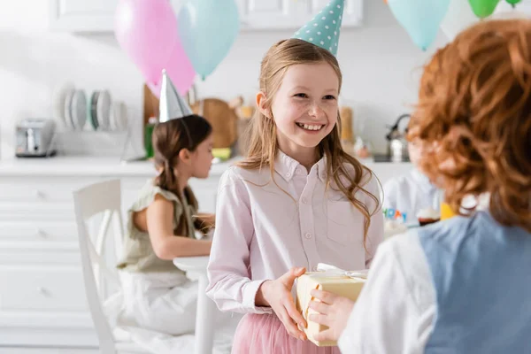 Happy girl in cap receiving present from redhead friend during birthday party — Stock Photo