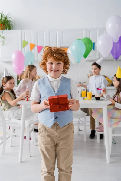 Happy redhead boy holding birthday present near friends during party at home — Stock Photo