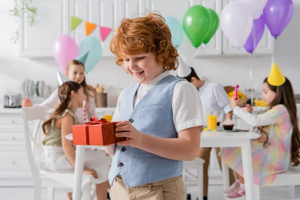 Веселий рудий хлопчик тримає подарунок на день народження біля друзів під час вечірки вдома — стокове фото