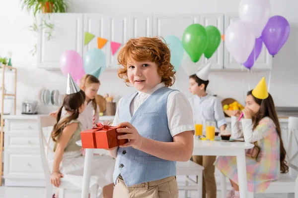 Кудрявый рыжий мальчик держит подарок на день рождения рядом с друзьями во время вечеринки дома — стоковое фото
