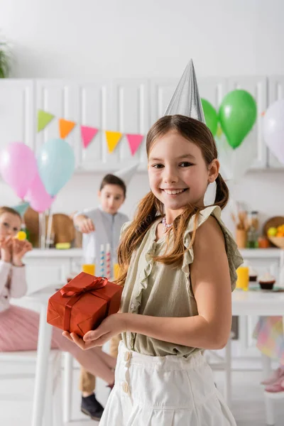 Щаслива дівчина на день народження у вечірній кепці тримає подарунок біля друзів на розмитому фоні — стокове фото
