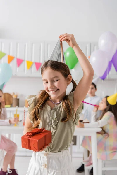 Menina alegre ajustando tampa do partido e segurando presente de aniversário perto de amigos no fundo borrado — Fotografia de Stock