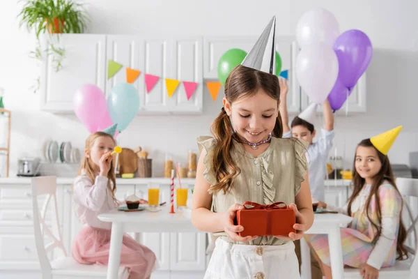 Pleased girl in party cap holding birthday present near friends on blurred background — Stock Photo