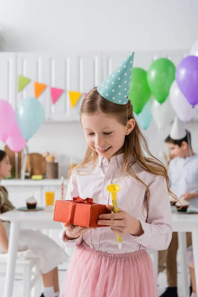 Happy preteen girl holding birthday present and party horn near friends on blurred background — Stock Photo