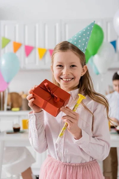 Cheerful preteen girl holding birthday present and party horn at home — Stock Photo