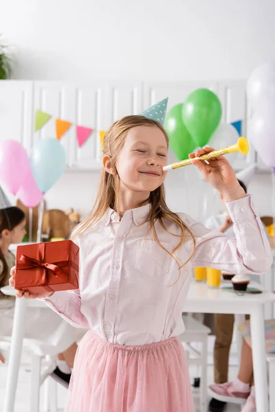 Happy preteen girl with closed eyes holding birthday present and party horn near friends on blurred background — Stock Photo