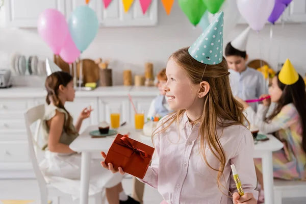Happy preteen girl holding birthday gift box and party horn near friends on blurred background — Stock Photo