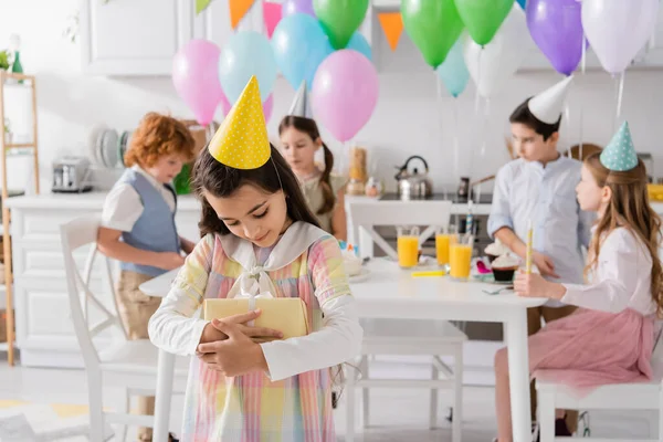 Счастливая девочка-подросток в шапочке для вечеринок обнимает подарок на день рождения рядом с друзьями на размытом фоне — стоковое фото