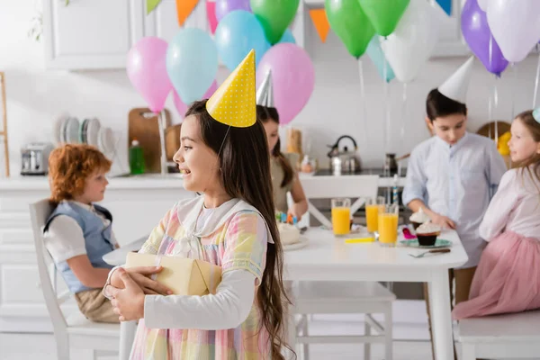 Cheerful birthday girl in party cap holding gift box near friends on blurred background — Stock Photo
