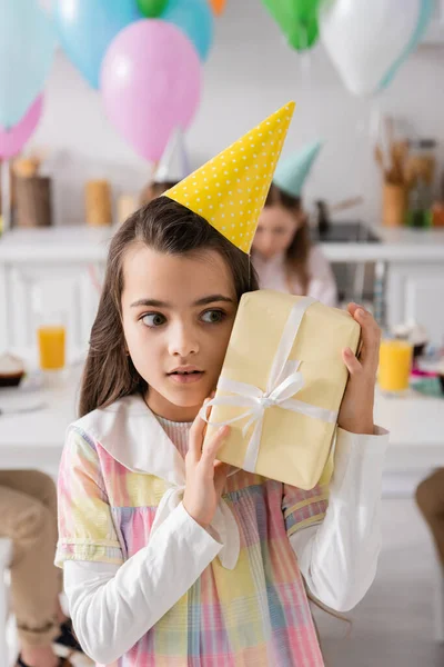 Stunned birthday girl in party cap holding gift box near friends on blurred background — Stock Photo