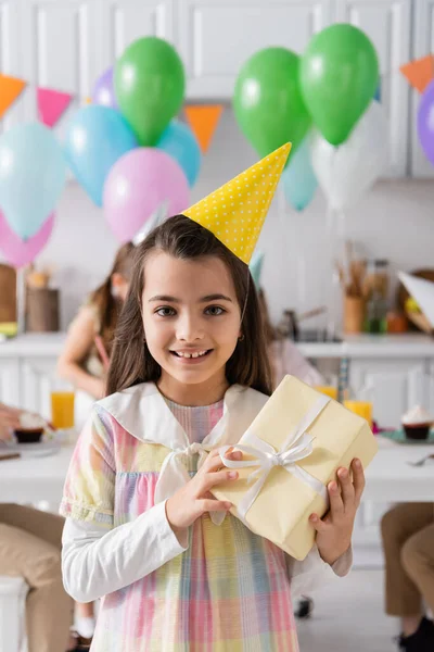 Cheerful birthday girl in party cap holding wrapped gift box near friends on blurred background — Stock Photo