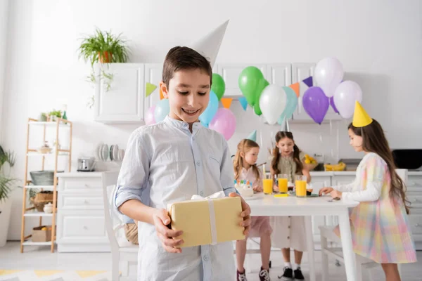 Menino pré-adolescente feliz em aparelho segurando presente de aniversário perto de amigos no fundo borrado — Fotografia de Stock