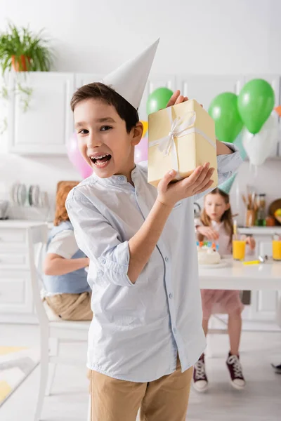 Збуджений хлопчик у дужках, що тримає подарунок на день народження біля друзів на розмитому фоні — стокове фото