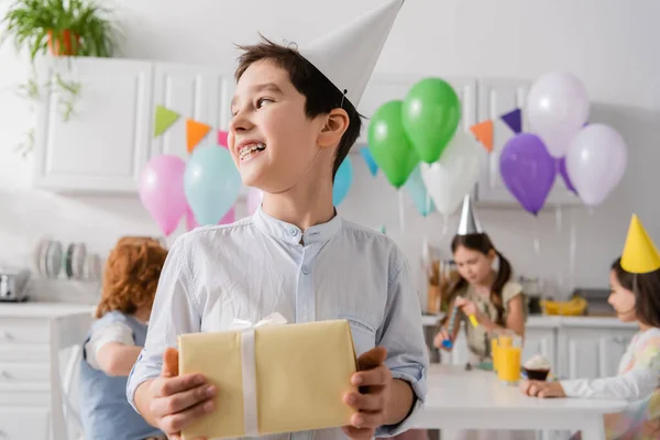 Cheerful preteen boy in braces holding birthday present near friends on blurred background — Stock Photo