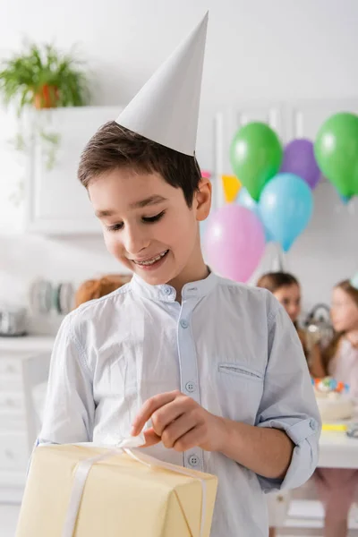 Sorrindo menino pré-adolescente em aparelho segurando presente de aniversário perto de amigos no fundo borrado — Fotografia de Stock