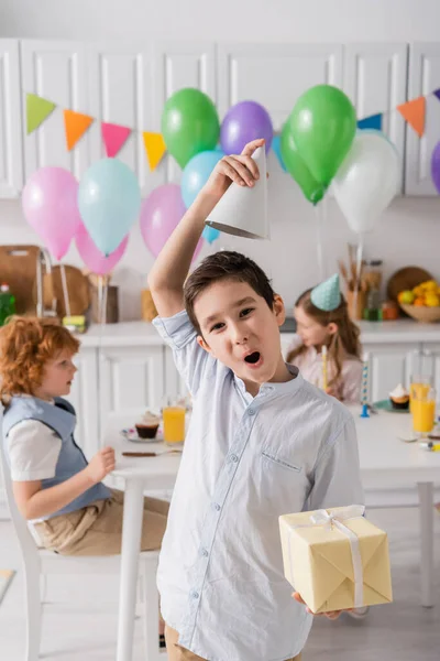 Funny preteen boy in braces holding birthday present and adjusting party cap near friends on blurred background — Stock Photo