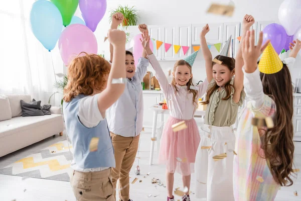 Cheerful kids in party caps dancing under falling confetti during birthday celebration at home — Stock Photo