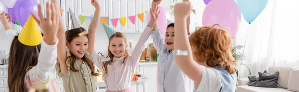 Счастливые дети в шапочках для вечеринок танцуют под падающими конфетти во время празднования дня рождения дома, баннер — стоковое фото