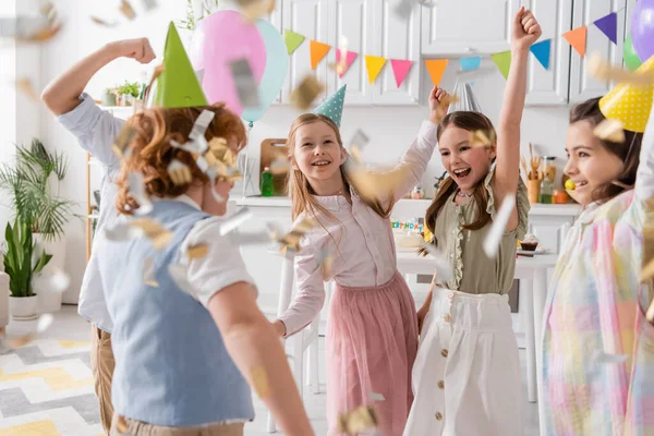 Grupo de crianças felizes em bonés de festa dançando sob queda confetti durante a celebração de aniversário em casa — Fotografia de Stock