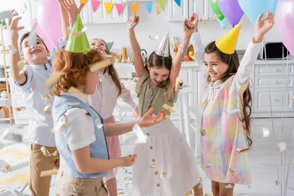 Grupo de niños alegres en gorras de fiesta bailando bajo confeti cayendo durante la celebración del cumpleaños en casa - foto de stock