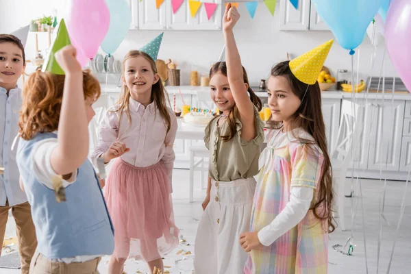 Группа счастливых детей в шапочках для вечеринок танцует во время празднования дня рождения дома — стоковое фото