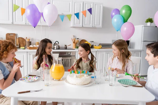 Grupo de niños alegres sosteniendo cuernos de fiesta y divirtiéndose durante la fiesta de cumpleaños junto a la torta con velas - foto de stock