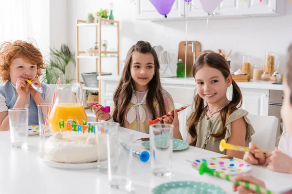 Happy girls and boy holding party horns next to birthday cake and looking at friend on blurred foreground — Stock Photo