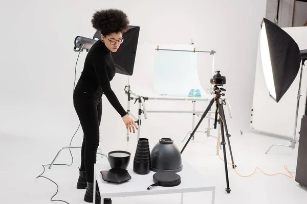 Élégant fabricant de contenu afro-américain atteignant l'équipement d'éclairage près du réflecteur softbox et appareil photo numérique sur trépied dans le studio photo — Photo de stock